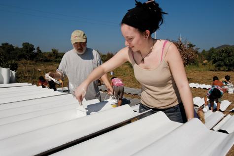 Two volunteers a man and a young woman ar up a roof painting it white. There are many other volunteers in the background.