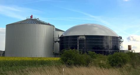 Three large containers at a biogas plant sit next to a field under a blue sky.