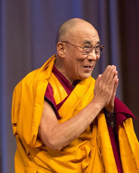 Image of the Dalai Lama standing in front of a purple curtain with his hands held in prayer. He is wearing traditional monks robes