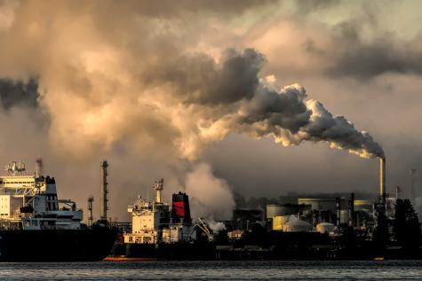 Image of large ships on the water in front of a huge power plant sending out large clouds of pollutant into the air 