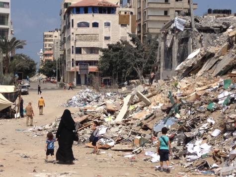 Image of people walking in the streets of Gaza with rubble all around them. There is a woman wearing a black burka holding the hand of a child in the foreground