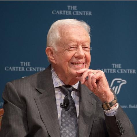 Photo of USA ex-president Jimmy Carter in his 90s. He is wearing a grey suit and smiling in front of a large sign for the Carter Center 