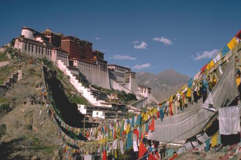 The Potala Palace in Lhasa, Tibetan Autonomous Region, China. A grand monastery looms in the background infront of a huge mountain range. Many prayer flags cover the valley