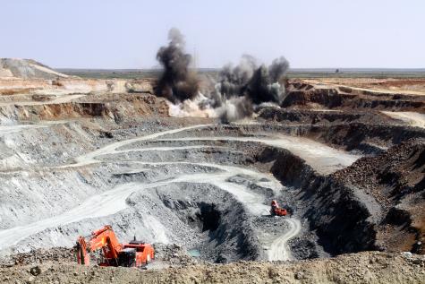 A huge open-pit mine shows the destruction of the mining industry, there is an explosion taking place in the background and machinery operating in the foreground.