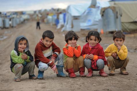 Five small children sit crouched down outside their refugee camp. They are wearing muddy wellies, you can see the white tents in the background.