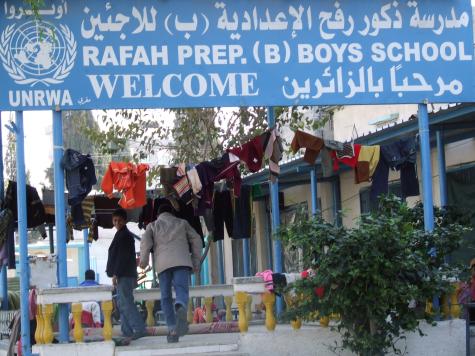 Image of a boys school in Rafah funded by UNRWA. A man and a boy are walking in the middle and there are lots of clothes hanging out to dry. There is a large blue sign at the entrance with the school's name on it. 