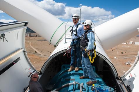 Two men wearing protective gear, helmets, and harnesses stand on top of a wind turbine which is open exposing the mechanics inside