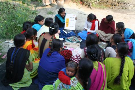 Many women sit on the floor taking part in a literacy class, they wear colourful dresses and there is reading material in the centre