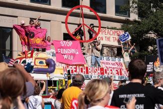 Colourful image of a bus decorated with peace symbols there are protesters riding on top displaying anti-war signs and against funding the war